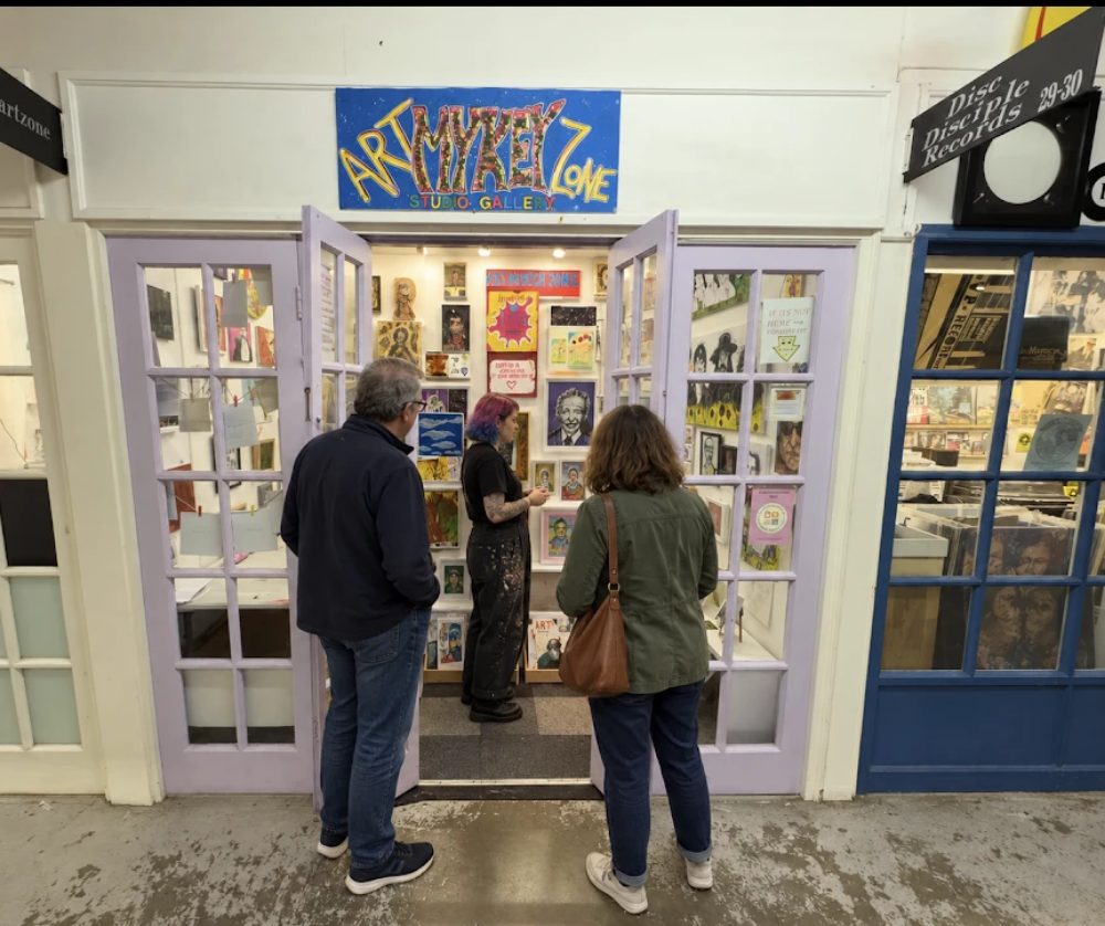 Mykeyartzone, a lilac double doorway under a hand-painted sign, the studio walls hung with prints and paintings and two visitors at the threshold